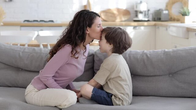 Loving mother at home with preschool child. Parent and kid bonding: happy mom kiss small son in forehead sit together with boy on sofa on weekend morning or quarantine lockdown. Family love concept