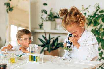 Obraz na płótnie Canvas Boy and girl in white uniforms and protective glasses do chemical experiments in a home laboratory.Back to school concept.Young scientists.Natural sciences.Preschool and school education of children.