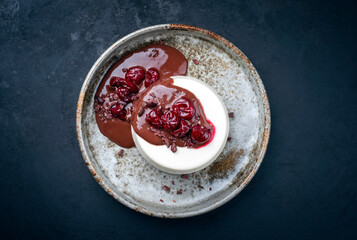 Modern style traditional blancmange almond pudding with cherry and chocolate cream and crumbles served as top view in a Nordic design plate on a black board with copy space