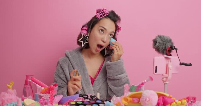 Professional female blogger records video on smartphone applies foundation on face with cosmetic sponge shoots makeup tutorial applies hair rollers makes hairstyle dressed in bathrobe poses at table