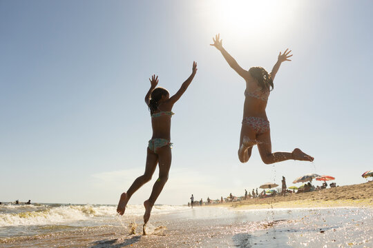 TWO CHEERFUL YOUNG GIRLS IN BIKINI JUMPING ON THE BEACH WITH OPEN ARMS. SUMMER HOLIDAYS CONCEPT, VACATIONS ON THE BEACH, LIFESTYLE, FREEDOM, JOY, FUN AND HAPPINESS.