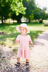 Little girl in a hat walks along the gravel path in the park with a stick in her hand