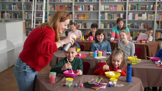 Friendly teacher squeezing shaving foam into bowls of little girl and boy sitting during master class for children with special needs. Kids with disabilities enjoying hobby