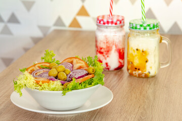 Bowl with escarole salad, red onion slices, olives stuffed with anchovies and tomatoes and fruit smoothies on the restaurant table in the background