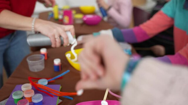 Hand of woman teacher squeezing shaving foam into bowls standing in front of kids with special needs. Children with disabilities making slimes with help of teacher during inclusive master class