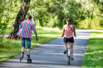Two friends riding a bicycle and roller skating in a park in summer.