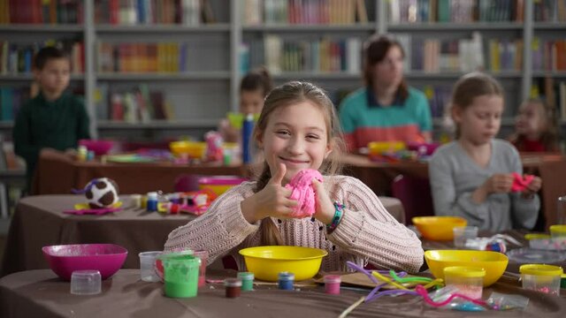 Smiling preteen child with special needs playing slime while posing for video sitting at table during masterclass. Cute girl with psychosyndrome posing on camera with self-made slime
