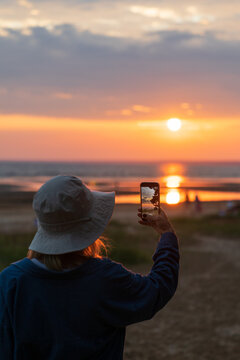 Silhouette of woman traveler wearing panama hat taking photo of golden sunset on smartphone, standing on the beach of sea or lake, rear view. 