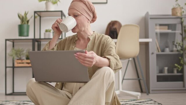 Tilting-up slowmo shot of happy mature woman in headscarf having cancer rehabilitation at home, drinking tea and using laptop sitting on floor in cozy living room