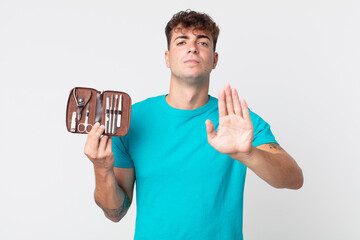 young handsome man looking serious showing open palm making stop gesture and holding a nails tools...
