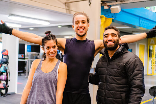 three friends in a gymnasium hugging posing for a photo, Latin woman and man and a Brazilian man, laughing