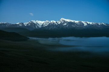 Atmospheric landscape with dense fog on lake and great snowy mountain ridge under night sky. Alpine scenery with thick fog on mountain lake and big mountain range in night. Snow pinnacle in dusk.