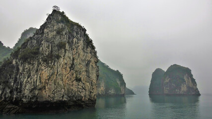 Fototapeta na wymiar Islands with bizarre outlines and steep slopes are hidden in the fog. Reflection on the surface of the calm water of Halong Bay. Vietnam