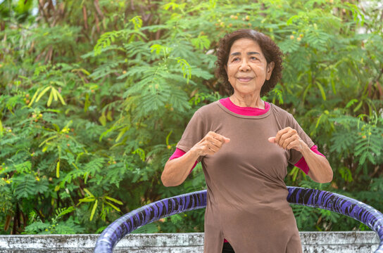 Portrait Of Healthy Asian Senior Woman Is Working Out By Hula Hooping On The Terrace Of House, Background Top Part Of Big Tree, Eyes Looking To The Blank Space For Copy And Design.