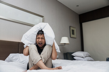 Sad asian boy covering his ears by pillow while screaming on the bed