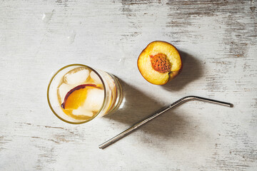 Peach refreshing beverage with ice, a half of fresh peach and metal straw for drinks on an old white wooden table, top view
