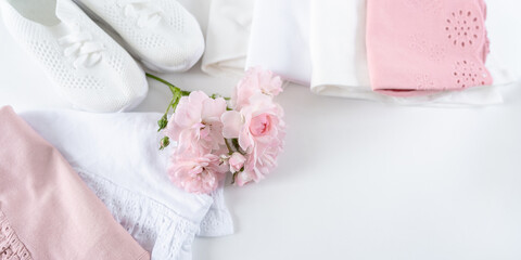 Set of white and pink baby clothes and shoes on white background with copy space decorated with fresh pink flowers