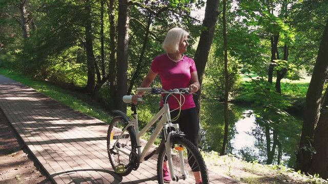 Mature sportive woman walks with her bike in a summer park. Concept of active lifestyle in nature, Elderly people healthcare, and healthy lifestyle.	