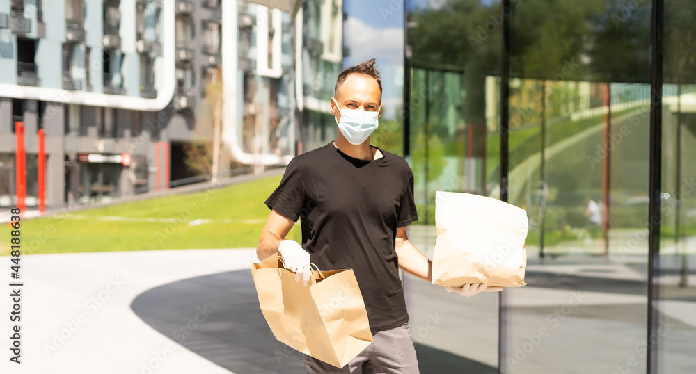 Wall mural Contactless delivery during COVID-19 pandemic lockdown concept. Courier wearing mask and gloves holds a parcel box and checking address details on the smartphone