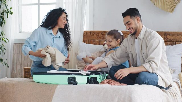 Multi-ethnic Family Multiracial Young Parents With Little Daughter Girl Child Sitting On Bed In Bedroom Packing Things In Suitcase Case Talking Getting Ready For Travel Trip Vacation Holiday Abroad