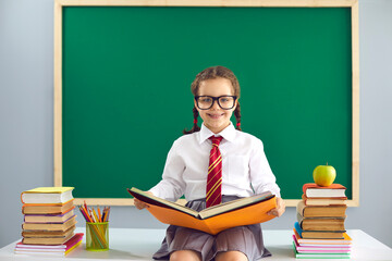 Back to school. A smart schoolgirl in glasses with a book in her hands is smiling on the background of the school blackboard.