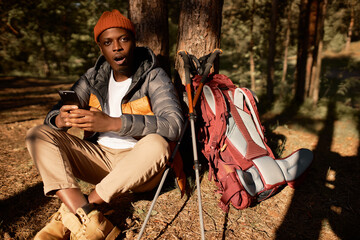 Shocked man saw bad news on smartphone. Shock and surprise, lost in forest, low internet connection, no navigation. Guy sit under tree with backpack and equipment. Travelling, nature, camping concept
