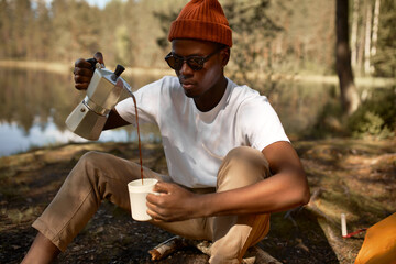 Young man pouring coffee in cup, morning breakfast on open air. Afro american traveler camping for...