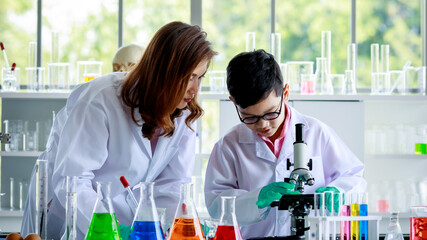 Concentrated Asian boy examining liquids through microscope while conducting experiment in school laboratory with female teacher.