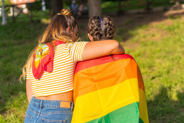 Closeup shot of two young Caucasian hugging females with LGBT pride flag sitting outdoors - obrazy, fototapety, plakaty