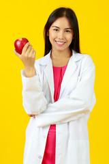 Asian young beautiful kind female doctor wearing white gown uniform with stethoscope, smiling and looking at red apple. Presenting nutrition, vitamin, healthy with isolated yellow background.