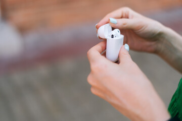 Close-up hands of unrecognizable young woman open white case with wireless earpiece standing in city street in summer day, blurred background, selective focus.