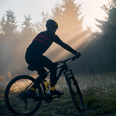 Silhouette of man in cycling suit riding bicycle in forest illuminated by morning sunlight. Male bicyclist cycling on grassy hill in the morning. Concept of sport, bicycling and active leisure.
