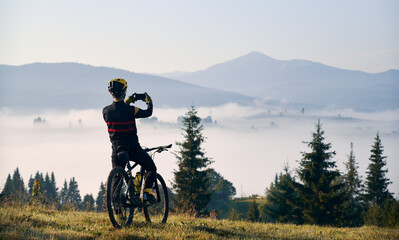 Fototapeta na wymiar Back view of young man in cycling suit sitting on bicycle and talking mountain photo with modern smartphone. Bicyclist in safety helmet holding cellphone and taking picture of beautiful misty hills.