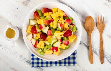 Mixed fruits salad including strawberry, kiwi, apple, and pineapple in white dish place on sackcloth on wooded table.  Utensil spoon and fork and oil salad dressing cup beside