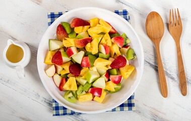 Mixed fruits salad including strawberry, kiwi, apple, and pineapple in white dish place on sackcloth on wooded table.  Utensil spoon and fork and oil salad dressing cup beside