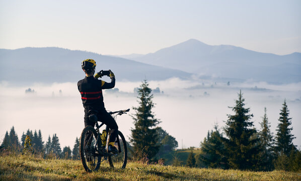 Back view of young man in cycling suit sitting on bicycle and talking mountain photo with modern smartphone. Bicyclist in safety helmet holding cellphone and taking picture of beautiful misty hills.