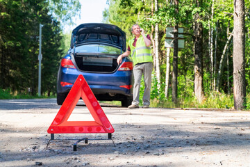 Close-up of a portable reflective red warning triangular sign on the side of a rural road near a car. The driver on the phone calling for technical assistance. Selective focus.
