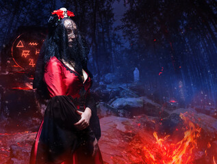 Mysterious witch in black veil, red gothic dress and crown with skull and roses standing in magical forest with fiery runes and ghosts. Halloween concept.