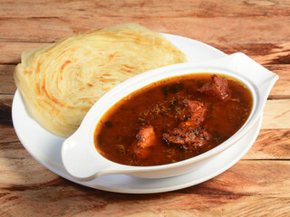 Kerala paratha / parotta, layered flat bread made from maida or whole wheat flour. Served with spicy kerala chicken curry, isolated over a rustic wooden table, selective focus