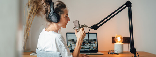 Wide image. Woman podcaster streaming and broadcasting her podcast from homemade studio. Female recording podcast from home and talking into a microphone.