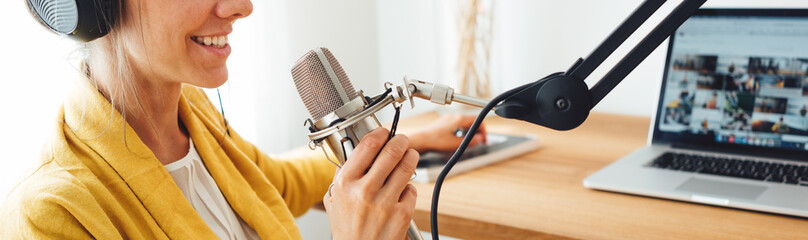 Wide image. Smiling female radio host recording and broadcasting her podcast from homemade studio. Woman talk into microphone on table - obrazy, fototapety, plakaty