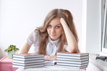 A girl with blue eyes and brown hair is sitting at a table, two stacks of books are in front of her, a girl is reading one book and, leaning her head on her hand, leans on her elbows