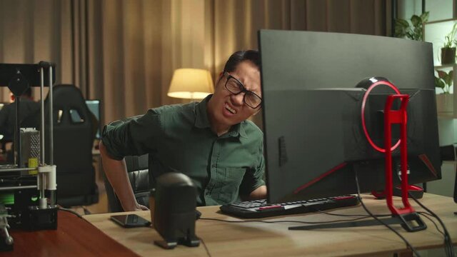Asian Man Paining His Back While Works On Personal Computer And 3D Printer In Home Office, He Works At Night 
