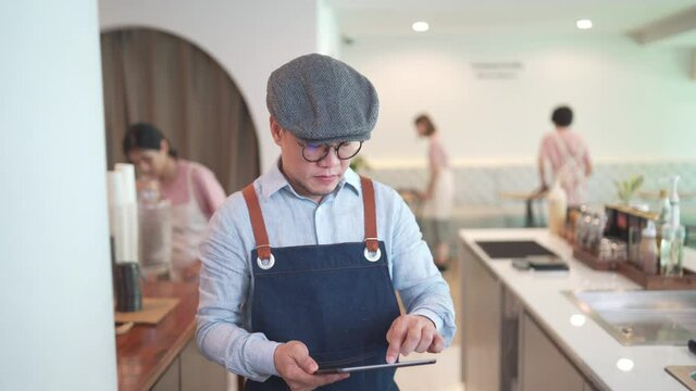 Asian man coffee shop manager walking behind bar counter working on digital tablet checking and counting inventory for ordering product for cafe. Small business restaurant owner entrepreneur concept