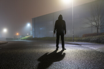 A hooded figure standing in the street in an empty industrial estate on a misty winters night.