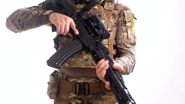 Man in combat camouflage and body armor attaches an ammunition magazine to his assault rifle and pulls the bolt into the firing position. White background. Isolated