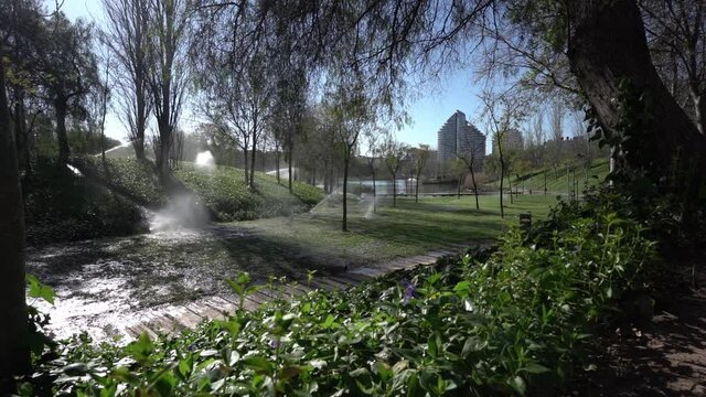 Static shot of a park with sprinkler irrigation system and buildings in the background