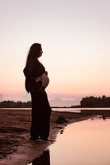 Pregnant woman on beach at sunset. Silhouette in profile against background of setting sun of beautiful young brunette with long hair and dark summer suit enjoying warm summer evening in nature. 