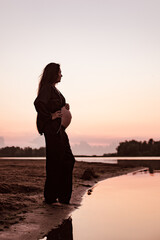 Fototapeta premium Pregnant woman on beach at sunset. Silhouette in profile against background of setting sun of beautiful young brunette with long hair and dark summer suit enjoying warm summer evening in nature. 