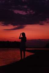 Fototapeta premium A slender woman is standing on the beach. A beautiful young happy woman admires the sunset and raises her hand to her head against the background of the river and picturesque clouds. 